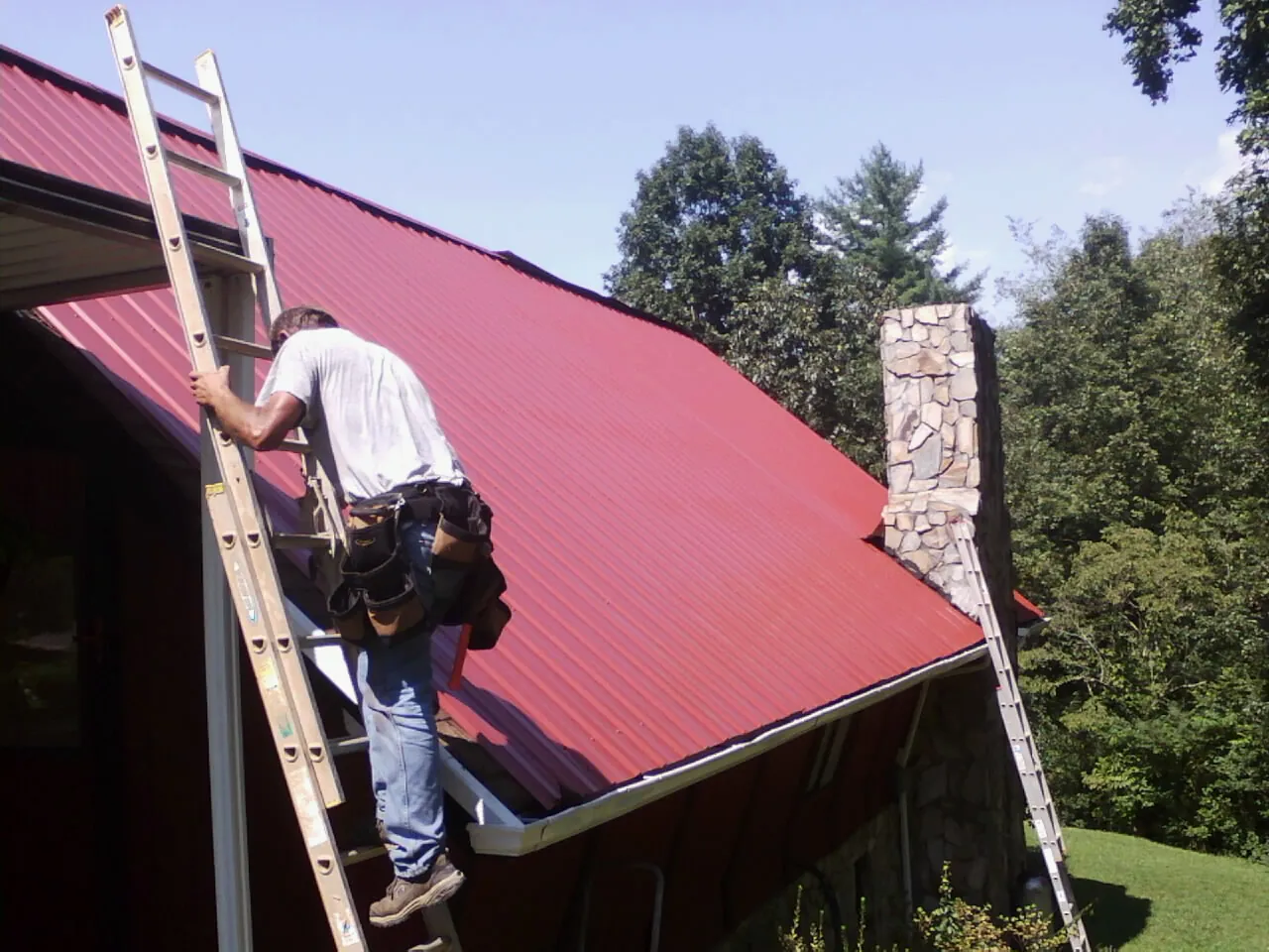 Roofer on a ladder beside a red metal roof and stone chimney — Soffit & Fascia Repair by Roofing Brothers in Norman